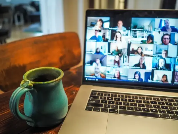 Laptop showing a video conference call on a wooden desk next to a teal mug